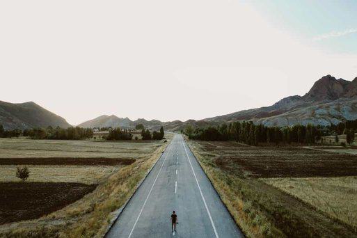 Aerial view of a two lane road cutting through farmland. There are hills and a person with short hair and a camera in the middle of the road faced off into the sunset. 