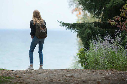 Woman left side of view with backpack looking onto horizon of water with trees on the right with bushes that have small purple flowers.